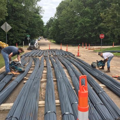 Workers cut rebar on a road; bundles of rebar are laid out. Cones and a stop sign are visible.