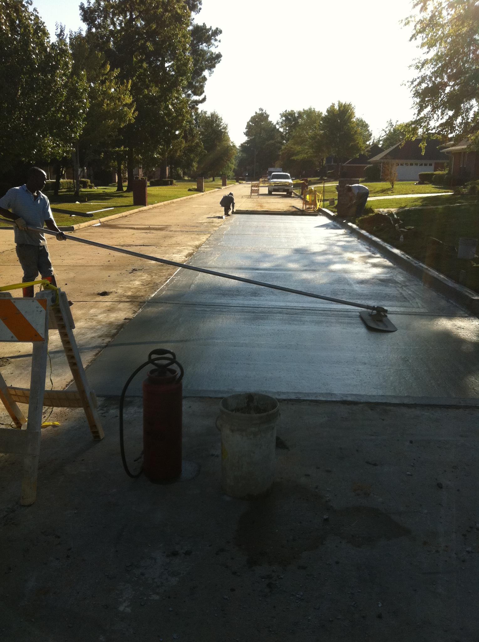 Worker smoothing fresh concrete on a street with a long handled tool.  Red fire extinguisher and bucket nearby.