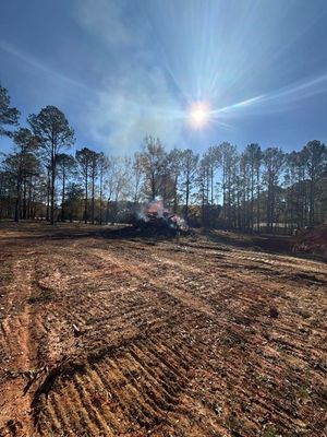 Dirt field with burning debris pile under bright sun, surrounded by trees.
