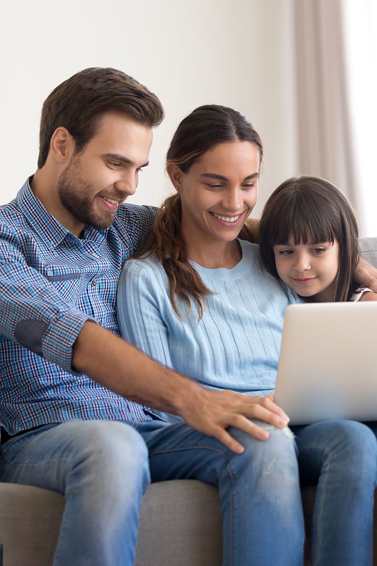 Family of three looking at a laptop together, smiling. Sitting on a couch in a well-lit living room.