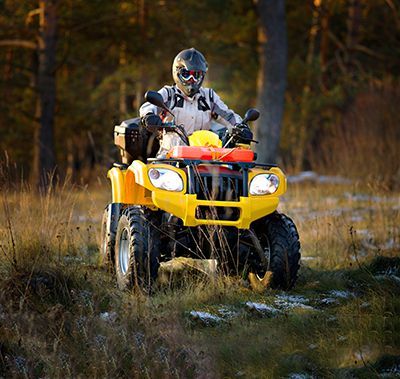 Person riding a yellow ATV in a grassy field. They wear a helmet and protective gear near a forest.