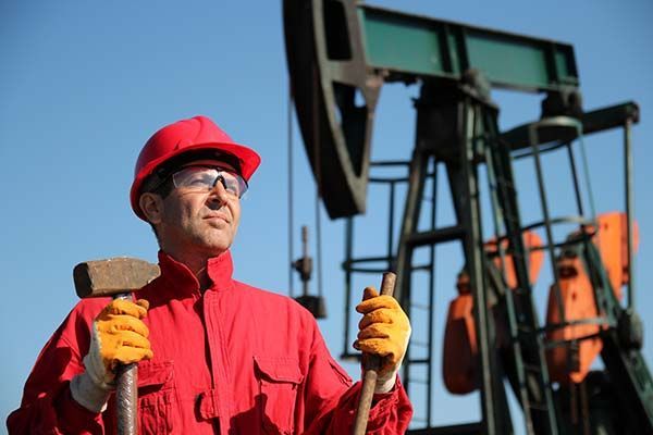 A man in a red hard hat is holding a hammer and shovel in front of an oil pump.