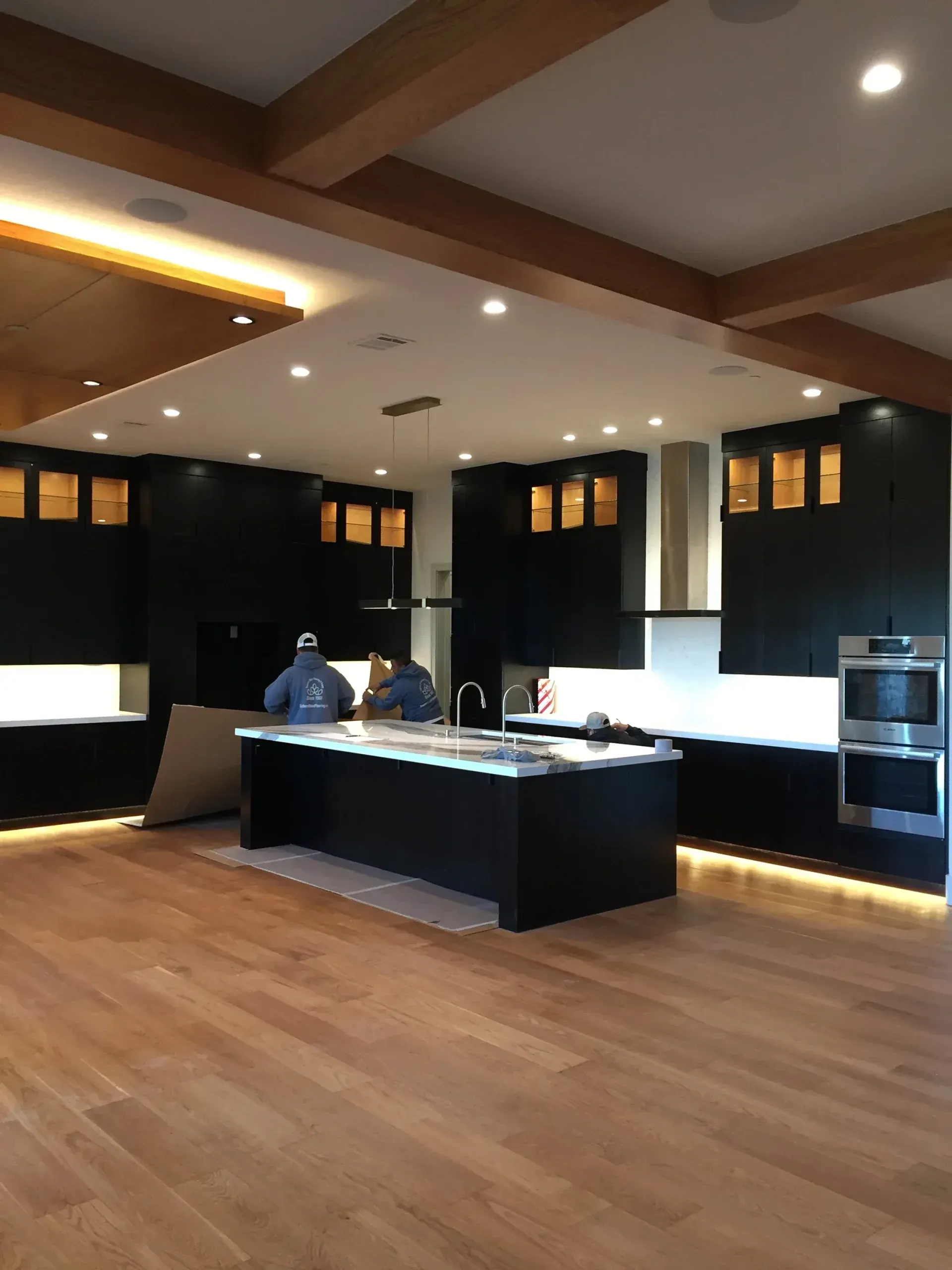 A modern kitchen with black cabinets, a large central island, light wood flooring, and recessed ceiling lighting.