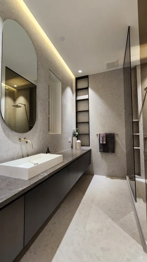 Modern bathroom with a long dark vanity, white rectangular sink, oval wall mirror, and a glass-enclosed shower area.