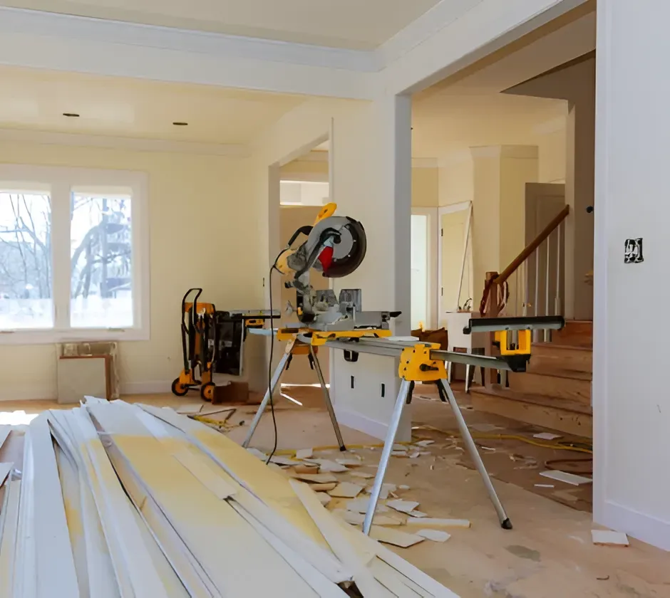 A miter saw on a stand sits in a room undergoing interior renovations with construction debris and trim on the floor.