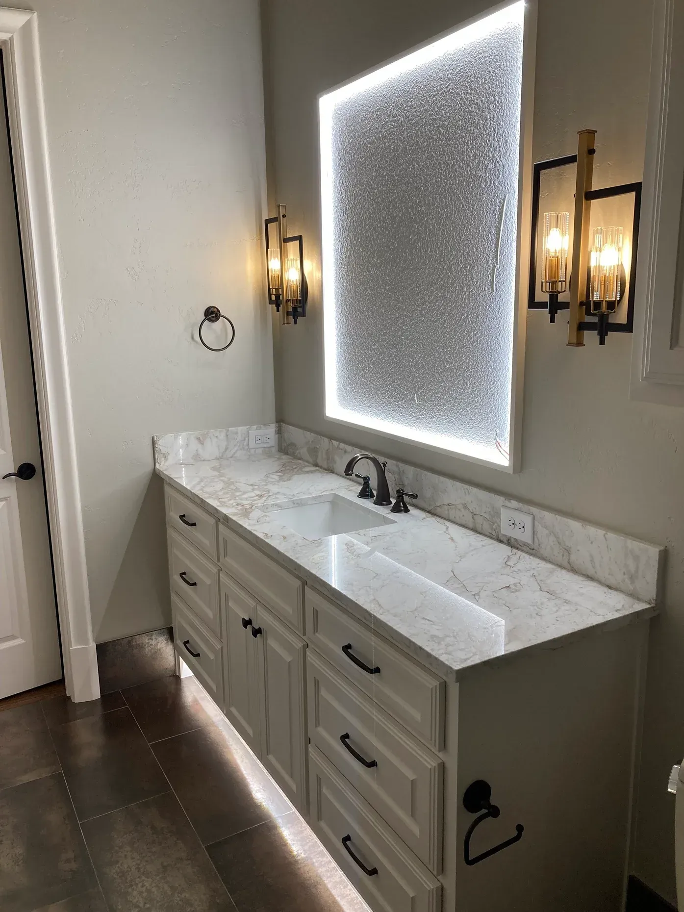 A modern bathroom vanity with white cabinets, a marbled countertop, a large lighted rectangular mirror, and wall sconces.