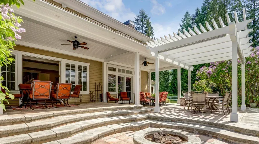 A patio with tiered stone steps, outdoor dining furniture, a pergola, and a central stone fire pit outside a house.