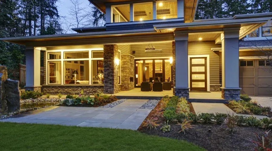 A modern two-story home at dusk with warm interior lighting, a stone pillar porch, a glass front door, and front lawn.