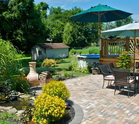 A backyard patio with pavers, a small pond, yellow flowering bushes, outdoor chairs, and two patio umbrellas.