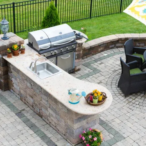 An outdoor kitchen with a stone base, a stainless steel grill, a sink, and a patio area with black chairs and a lawn.