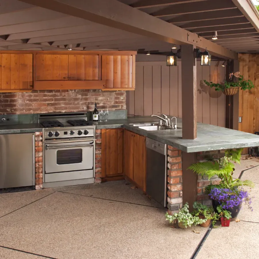 An outdoor kitchen with wood cabinets, brick walls, stainless appliances, and a slate countertop under a covered patio.