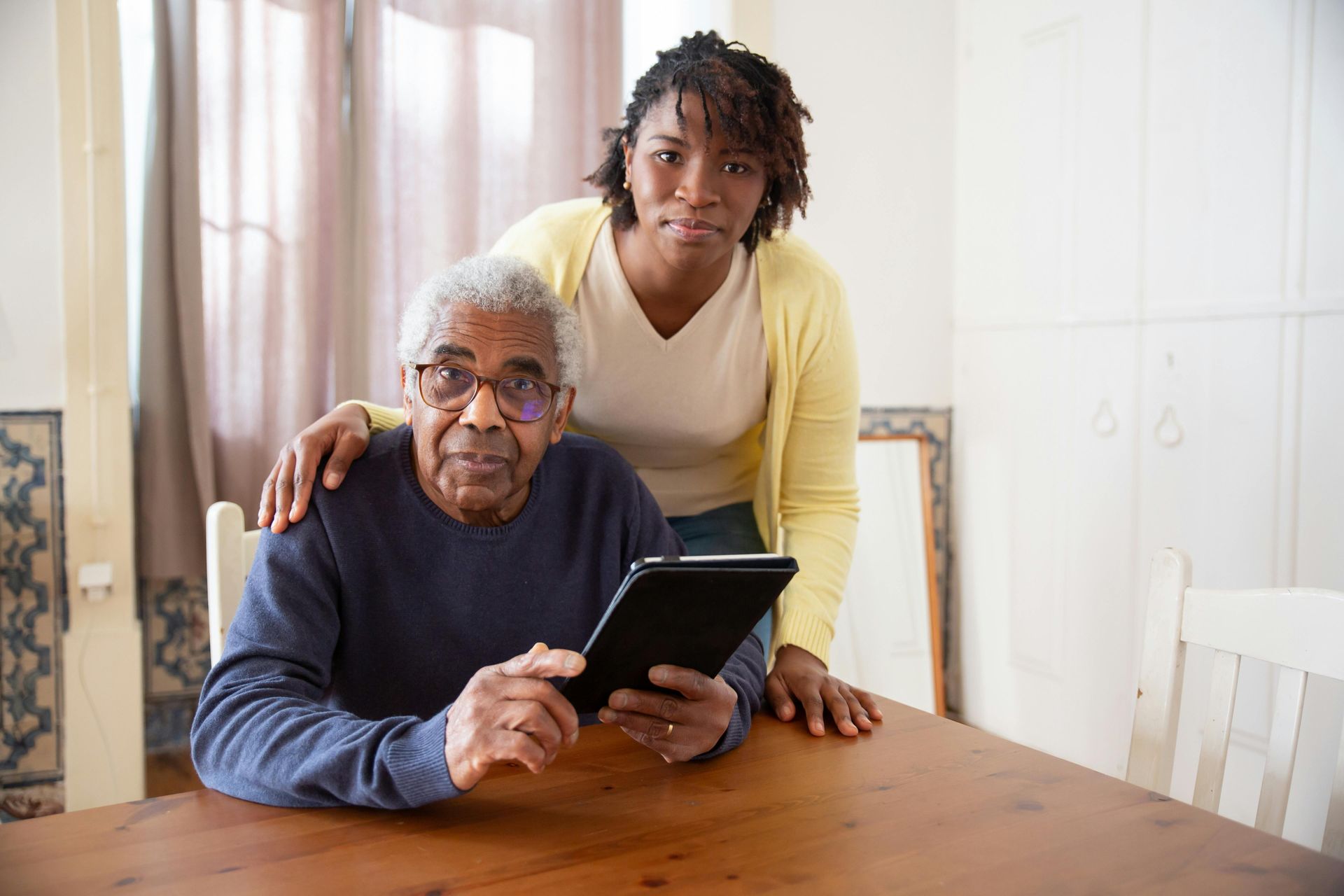An elderly man is sitting at a table using a tablet computer while a woman stands behind him.
