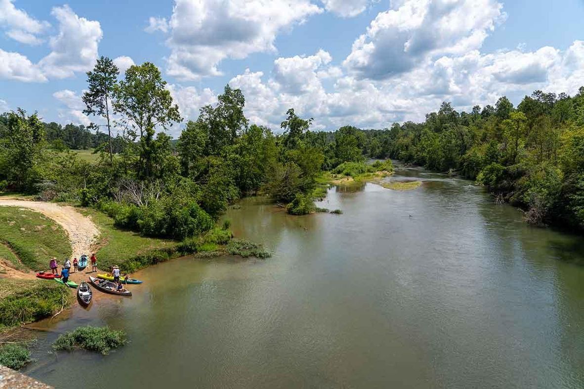People with kayaks and canoes gather on a grassy riverbank next to a calm, tree-lined river under a cloudy blue sky.