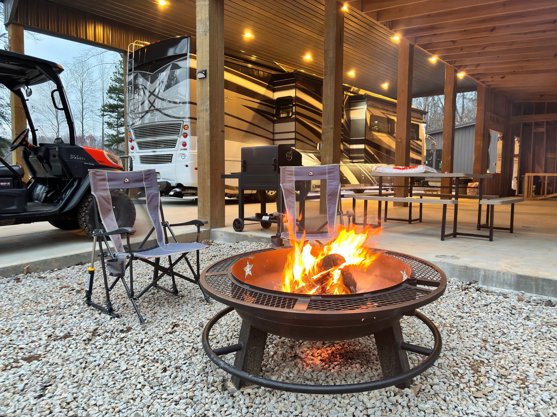 Fire pit with flames, chairs, and RV under a wooden canopy.