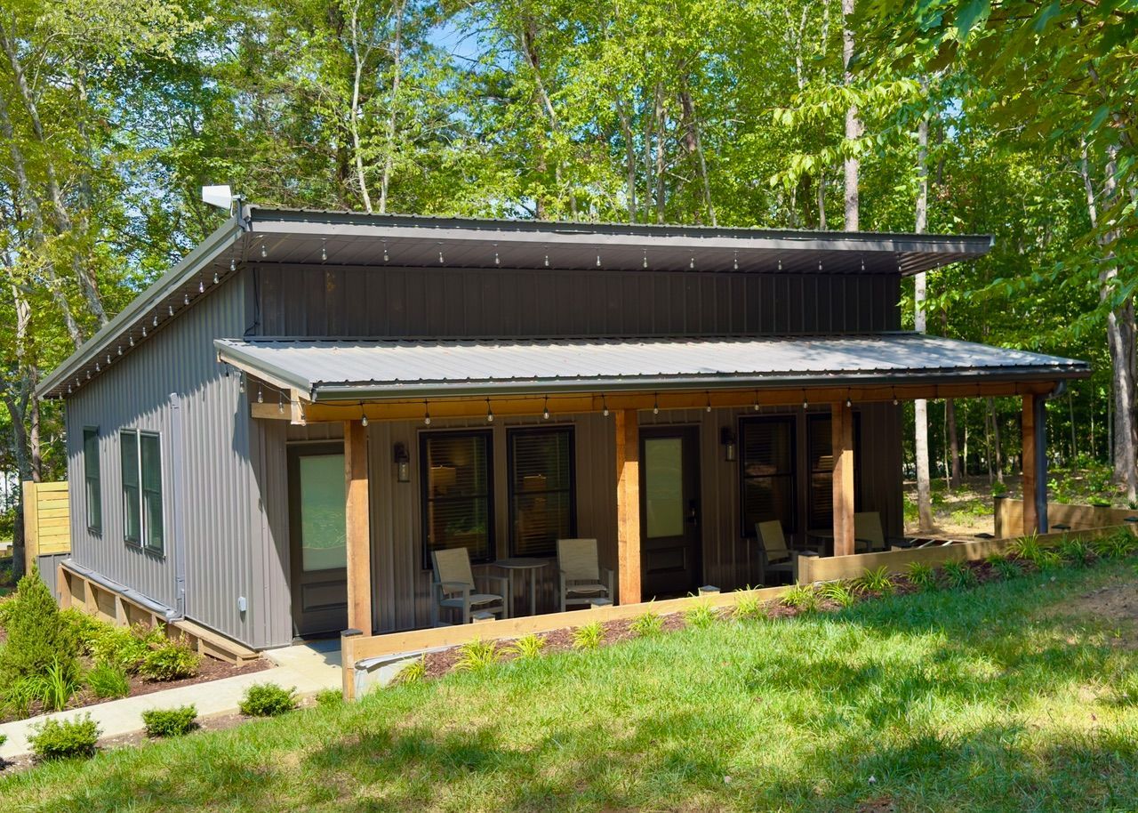 Modern charcoal-gray cabin with a metal roof and a wooden porch, nestled in a lush, sunny forest setting.