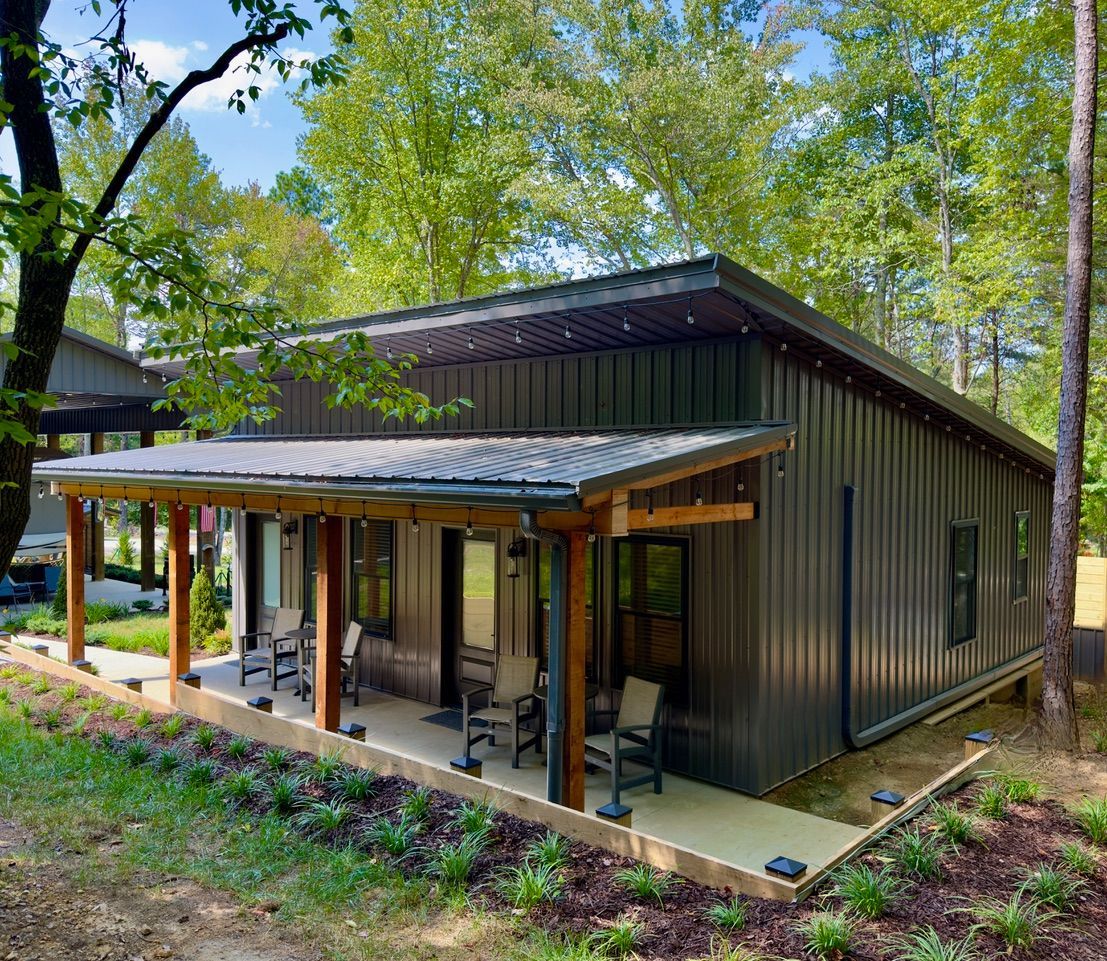 A modern, dark-sided cabin with a metal roof and a covered wooden porch, set in a wooded area with a landscaped border.