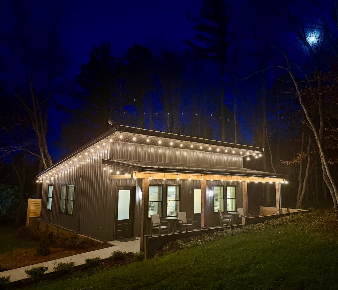 A dark-toned, single-story cabin illuminated by warm string lights on its porch at night, set against a forest backdrop.