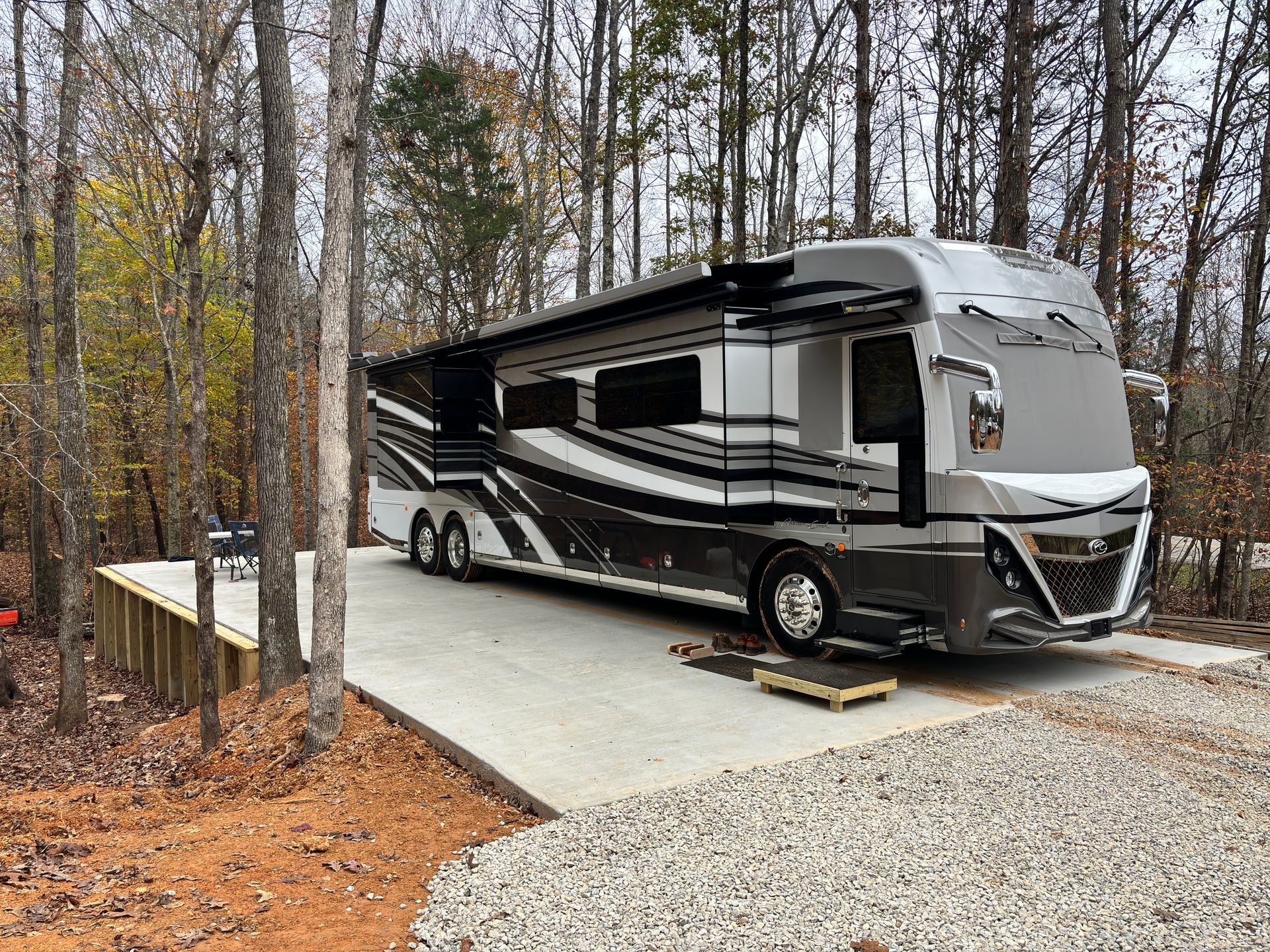 Large RV parked on a concrete pad in a wooded area, awning extended.