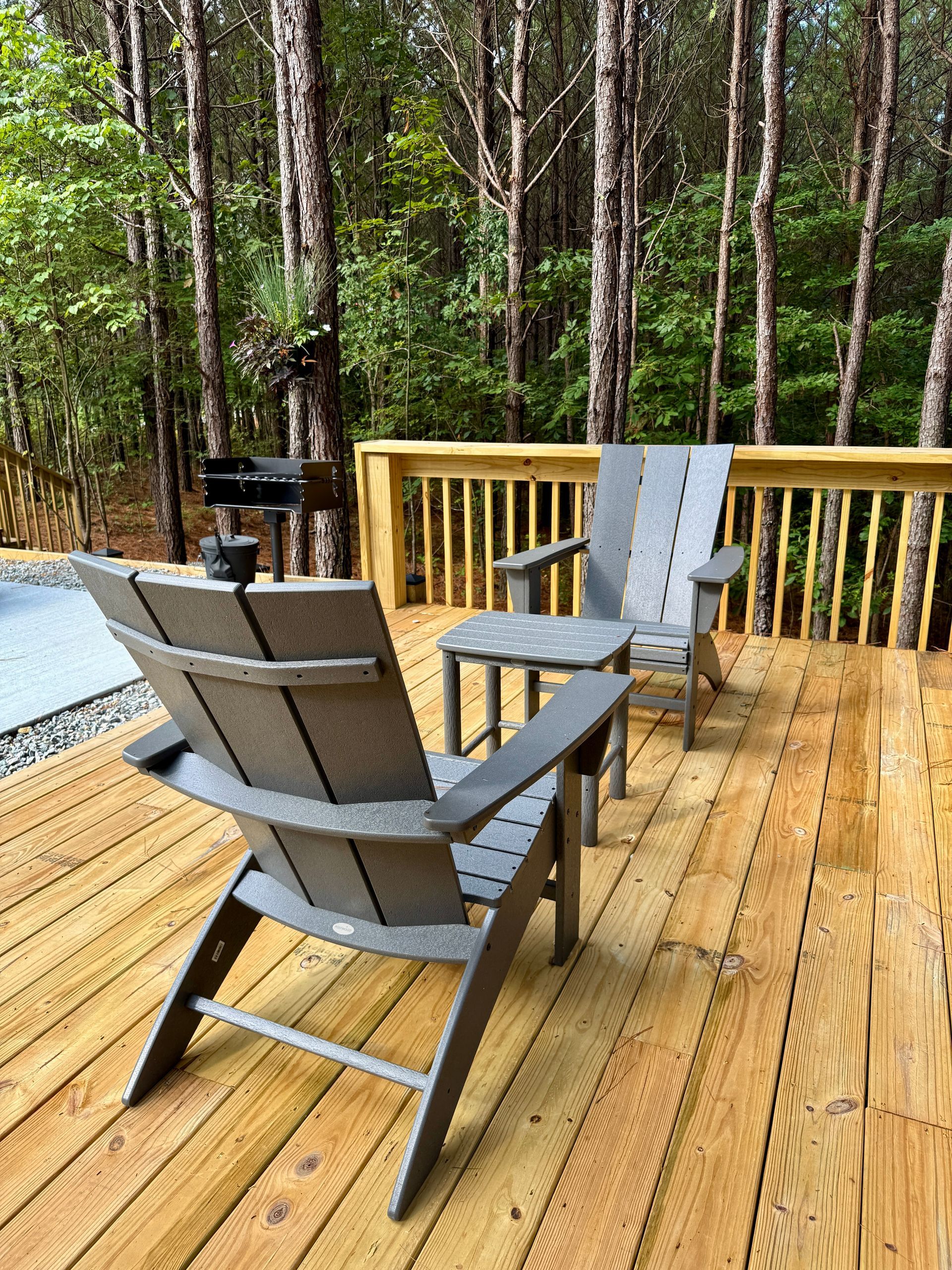 Two gray Adirondack chairs and a small table on a wooden deck in front of a forest.
