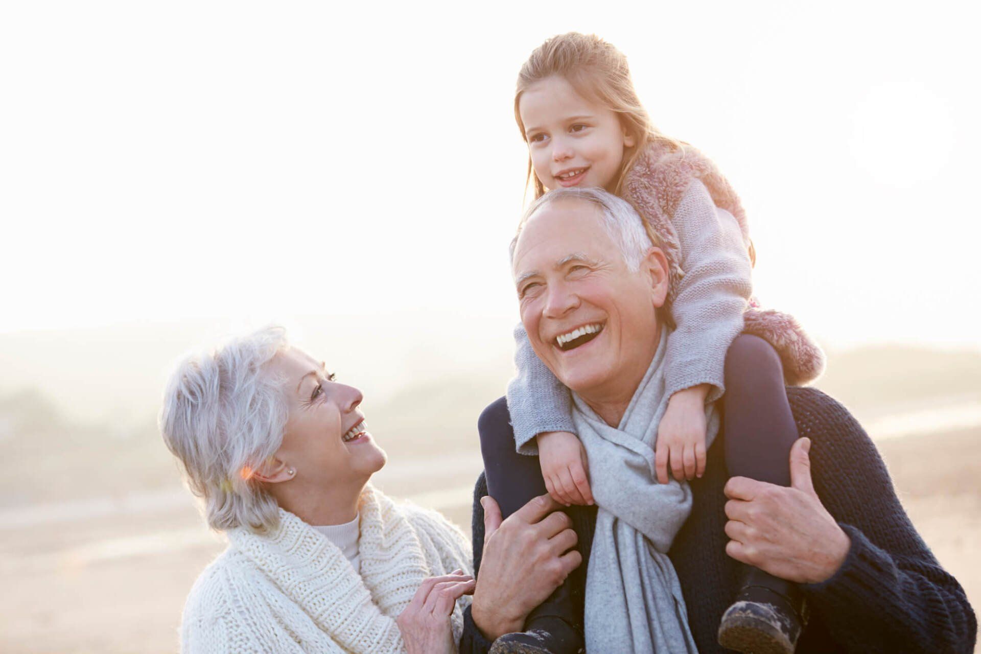 Grandparents and granddaughter on beach: man carries girl on shoulders, woman laughs, sunny background.
