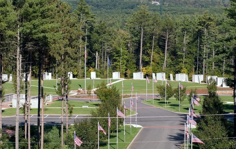 Cemetery with rows of white markers, American flags, and trees. Paved roads lead through the grounds.