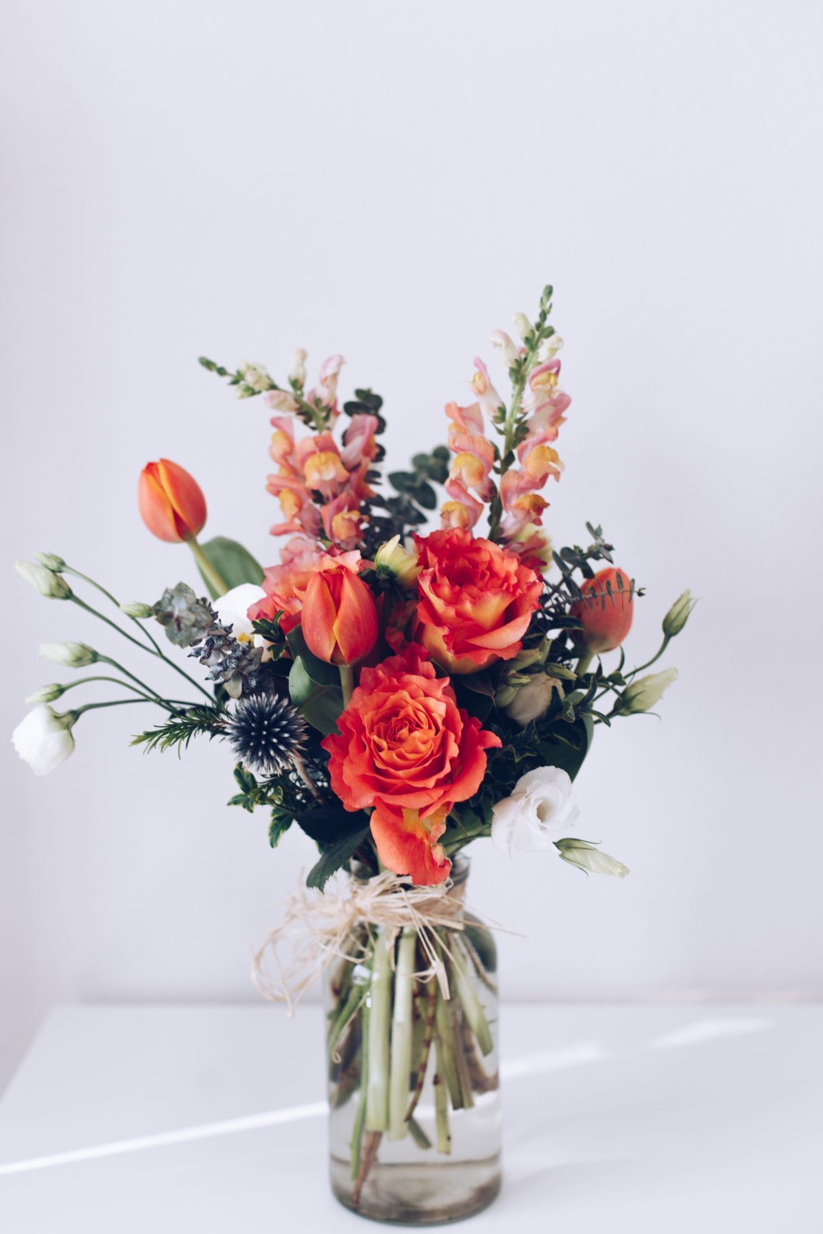 Bouquet of orange roses, tulips, and snapdragons in a glass vase on a white table.