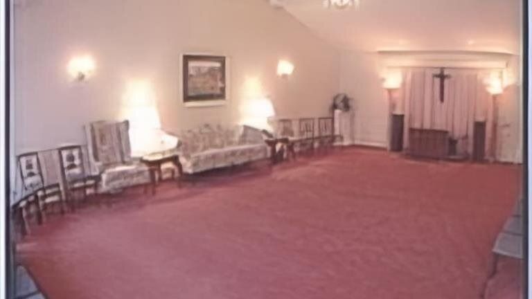 Interior of a funeral home chapel. Pink carpet, chairs, a sofa, and a cross at the altar.