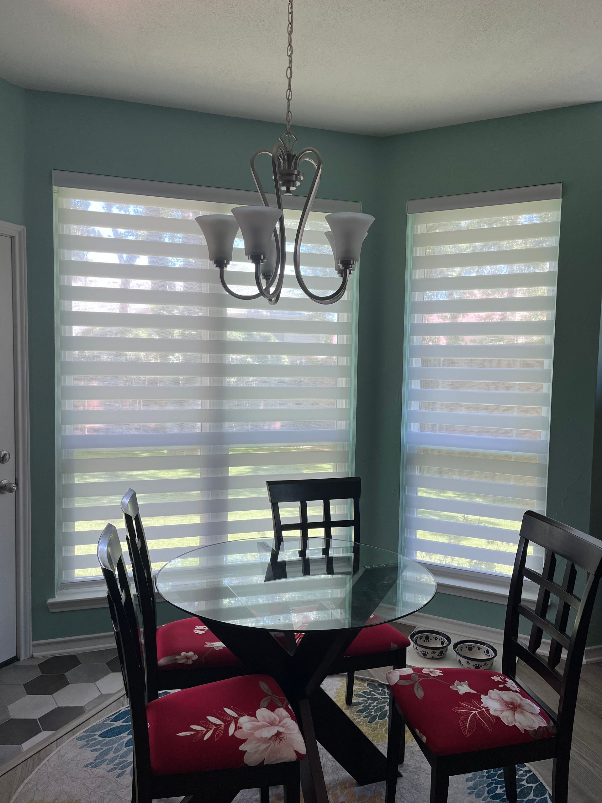 Dining room with round glass table, chairs, and layered blinds. Chandelier hangs above.