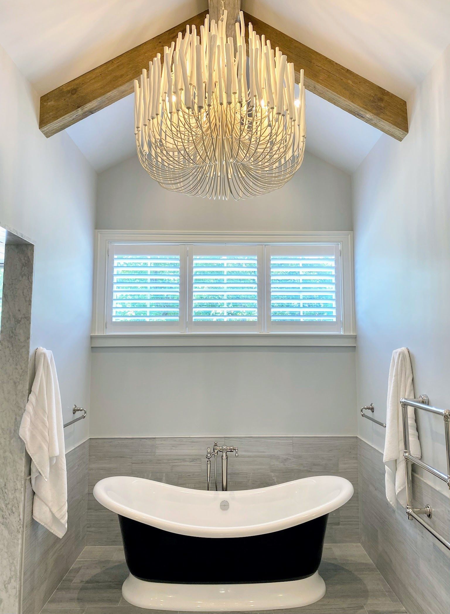 Bathroom with a black clawfoot tub, chandelier, and window with shutters. White and grey tones.