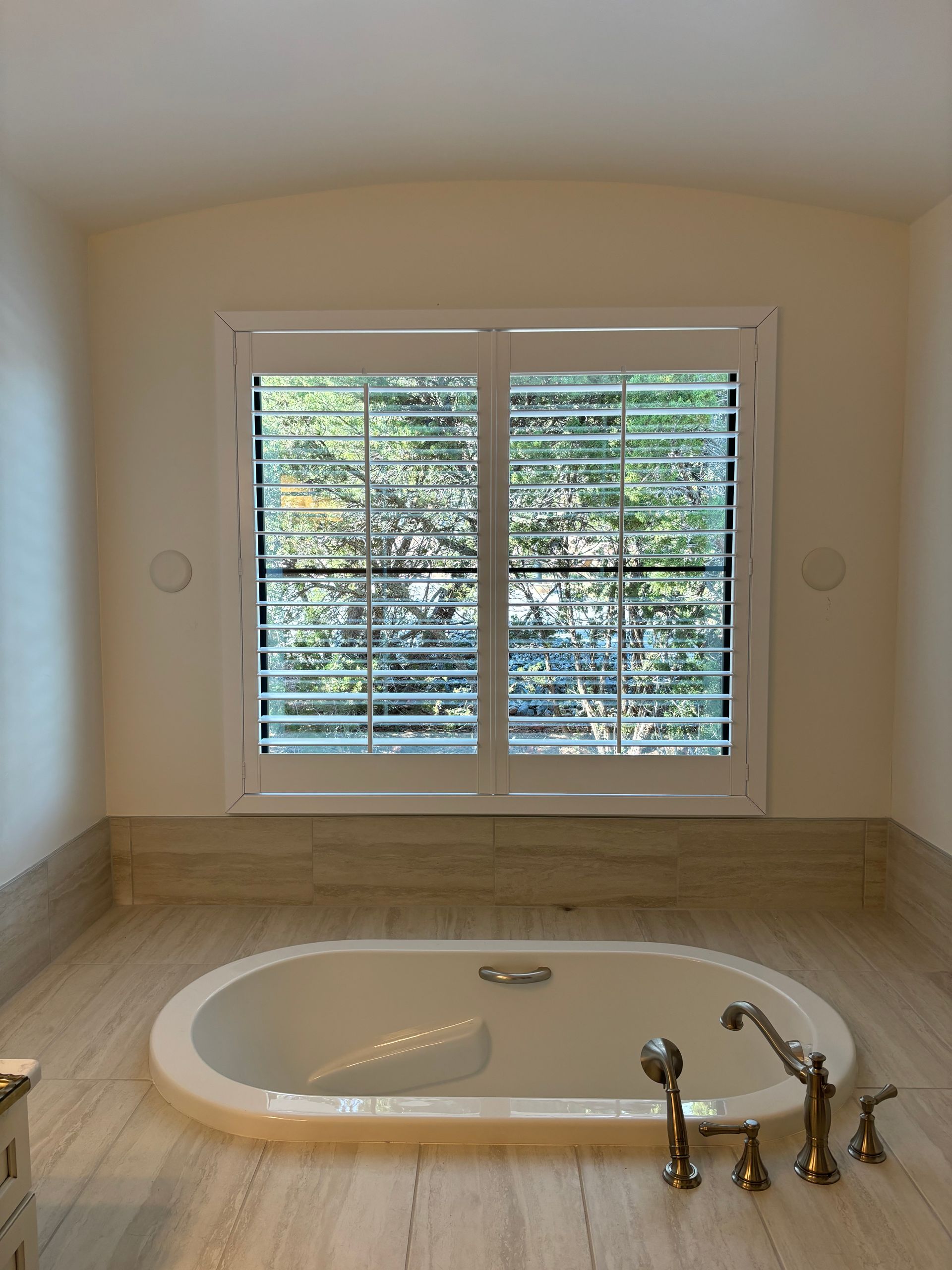 Bathroom with oval tub under a window with white shutters; light beige tile flooring.