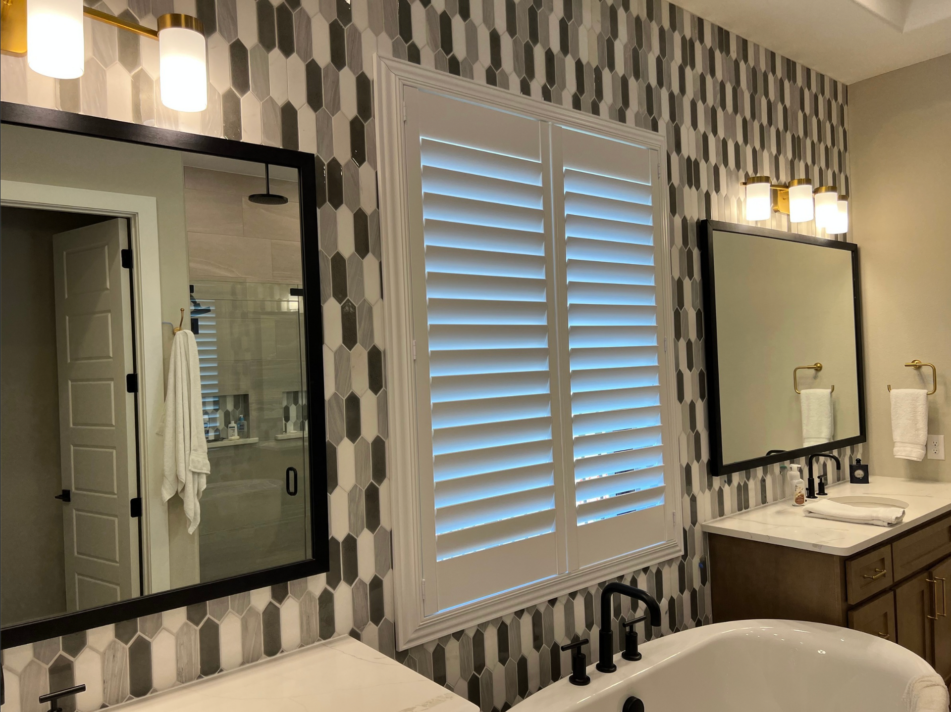Bathroom with dual sinks, patterned wall, tub, window shutters, and mirrors.