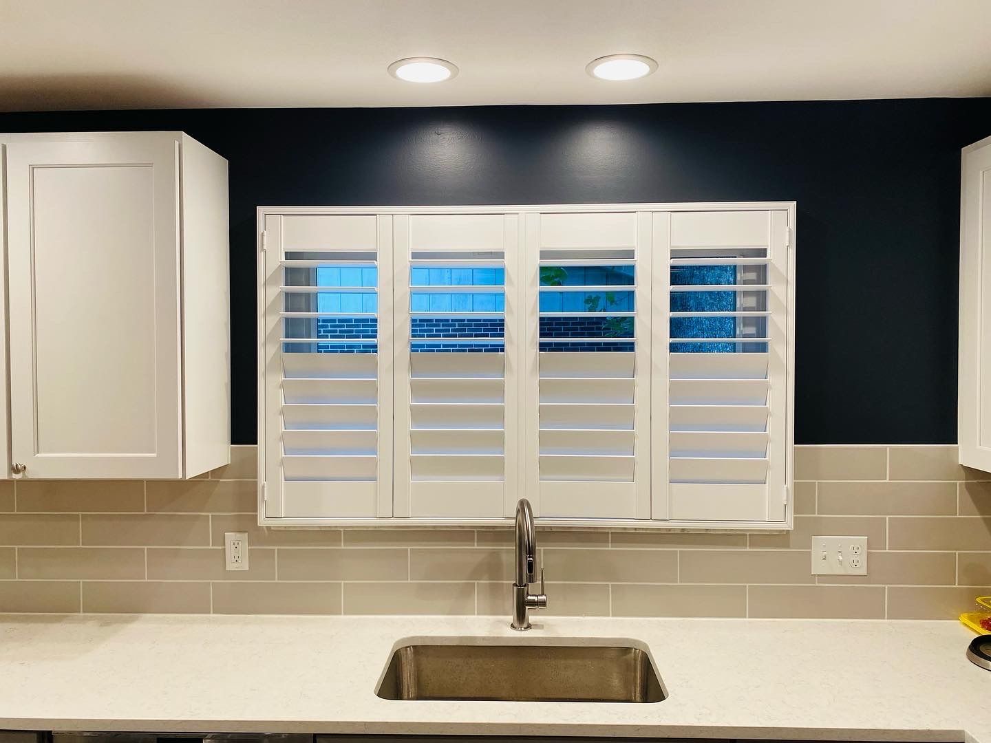 Kitchen sink with white shutters, cabinets, and backsplash against a dark blue wall.