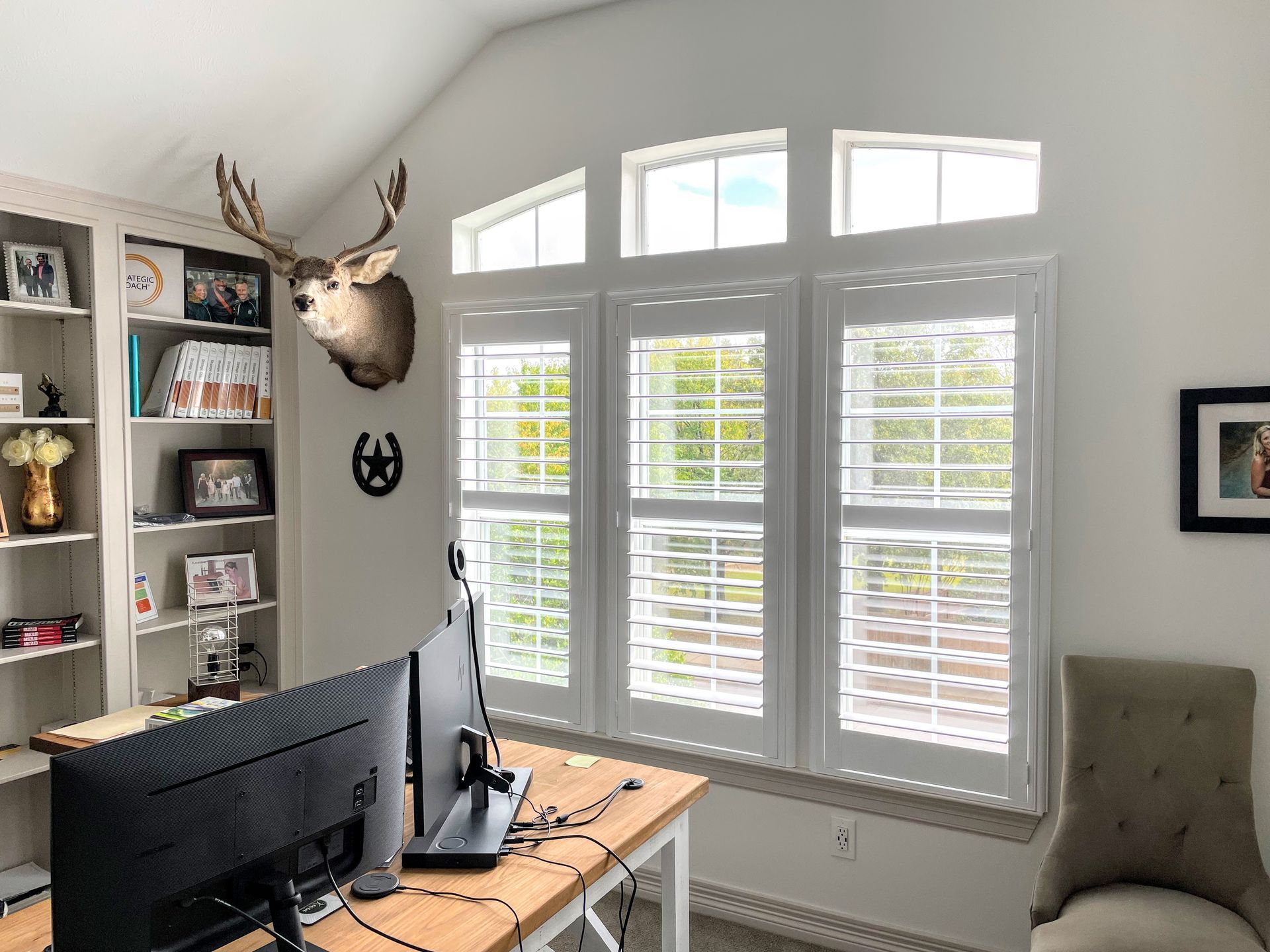 Office with white shutters, a desk with monitors, a bookcase, and a deer head mount.