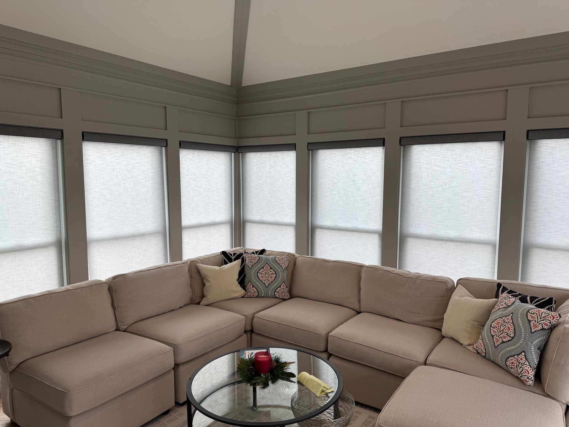 L-shaped beige sofa in a gray-walled room with roller shades. A glass coffee table holds holiday decor.