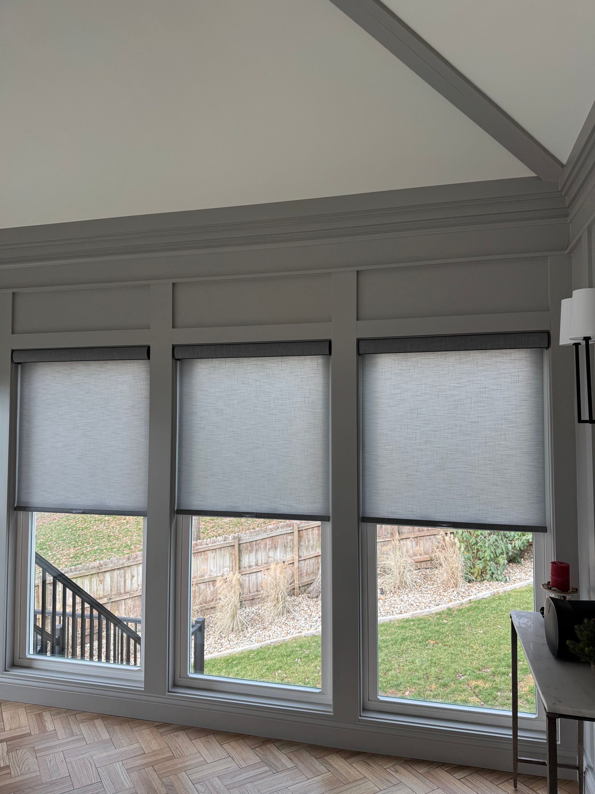 Three gray roller shades cover windows overlooking a green yard and black railing.