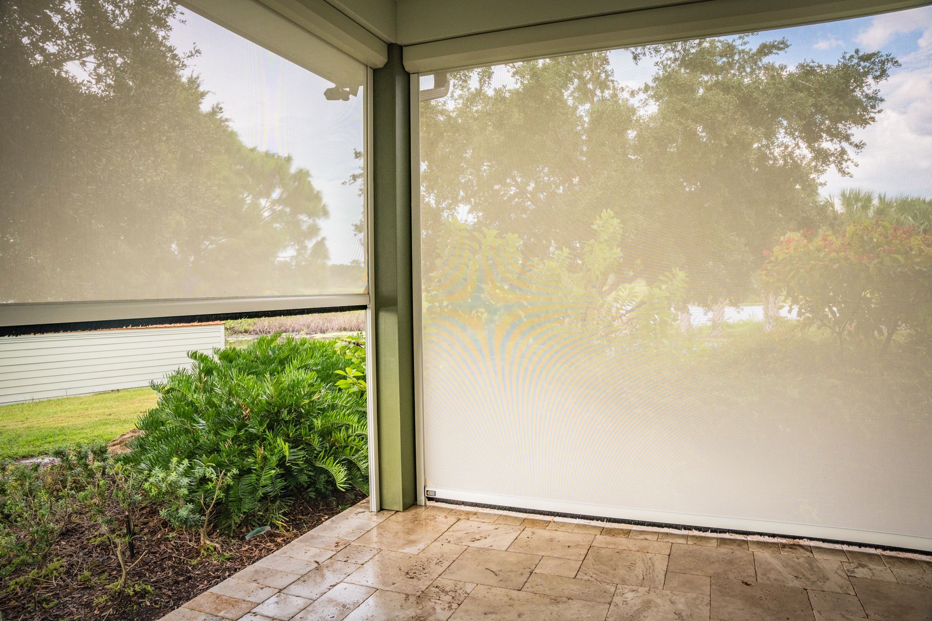 Outdoor patio with rolled-down beige solar shades, overlooking a lawn and trees.