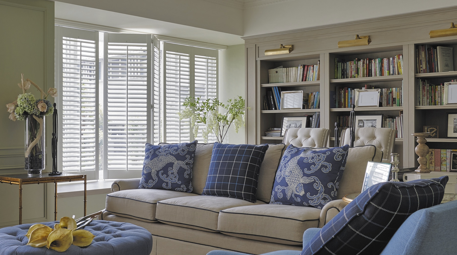 Cozy living room with a cream sofa, blue pillows, built-in bookshelves, and window with shutters.