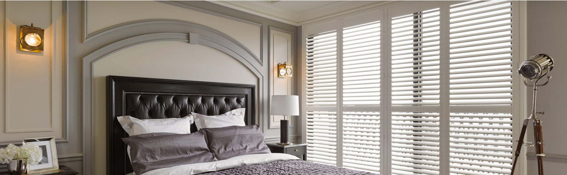 Bedroom with dark upholstered headboard, white shutters, and a tripod lamp. Soft lighting.
