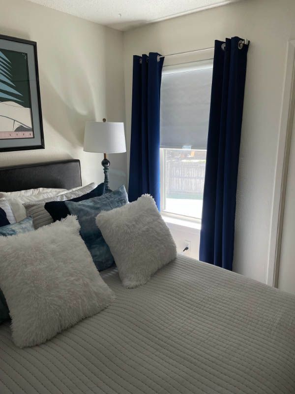 Bedroom with bed, pillows, blue curtains, lamp, and artwork.