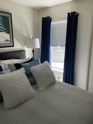 Bedroom with bed, blue curtains, lamp, art, and white fluffy pillows.