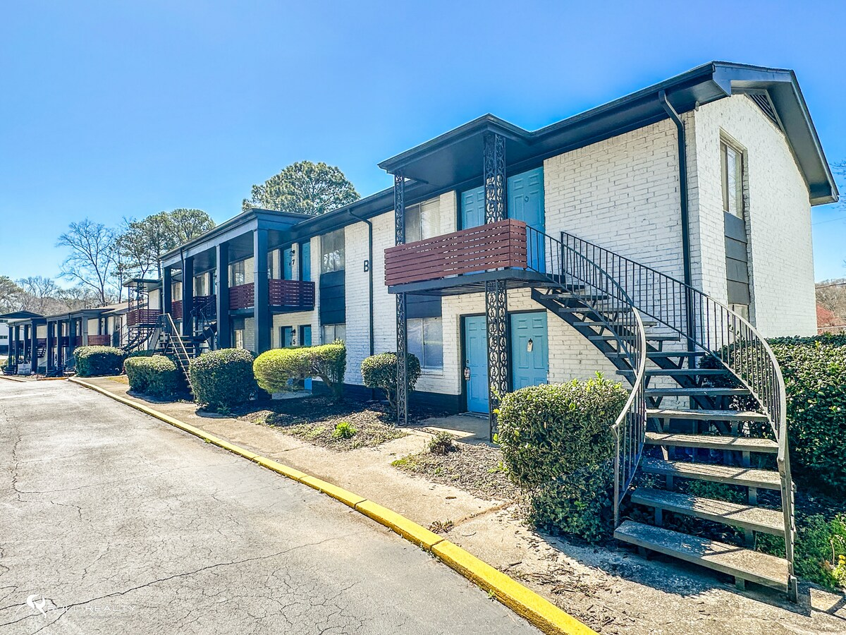 Apartment complex with exterior staircases, red balconies, and trimmed bushes on a sunny day