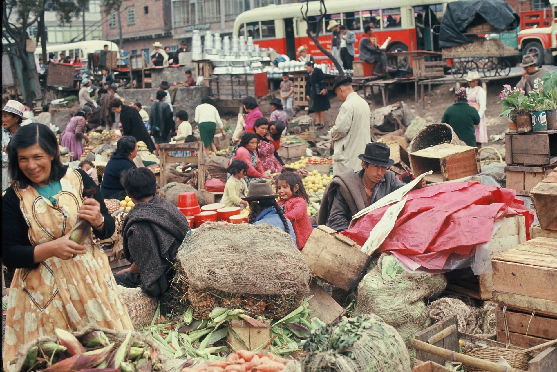 A group of people are gathered in a market with a red bus in the background