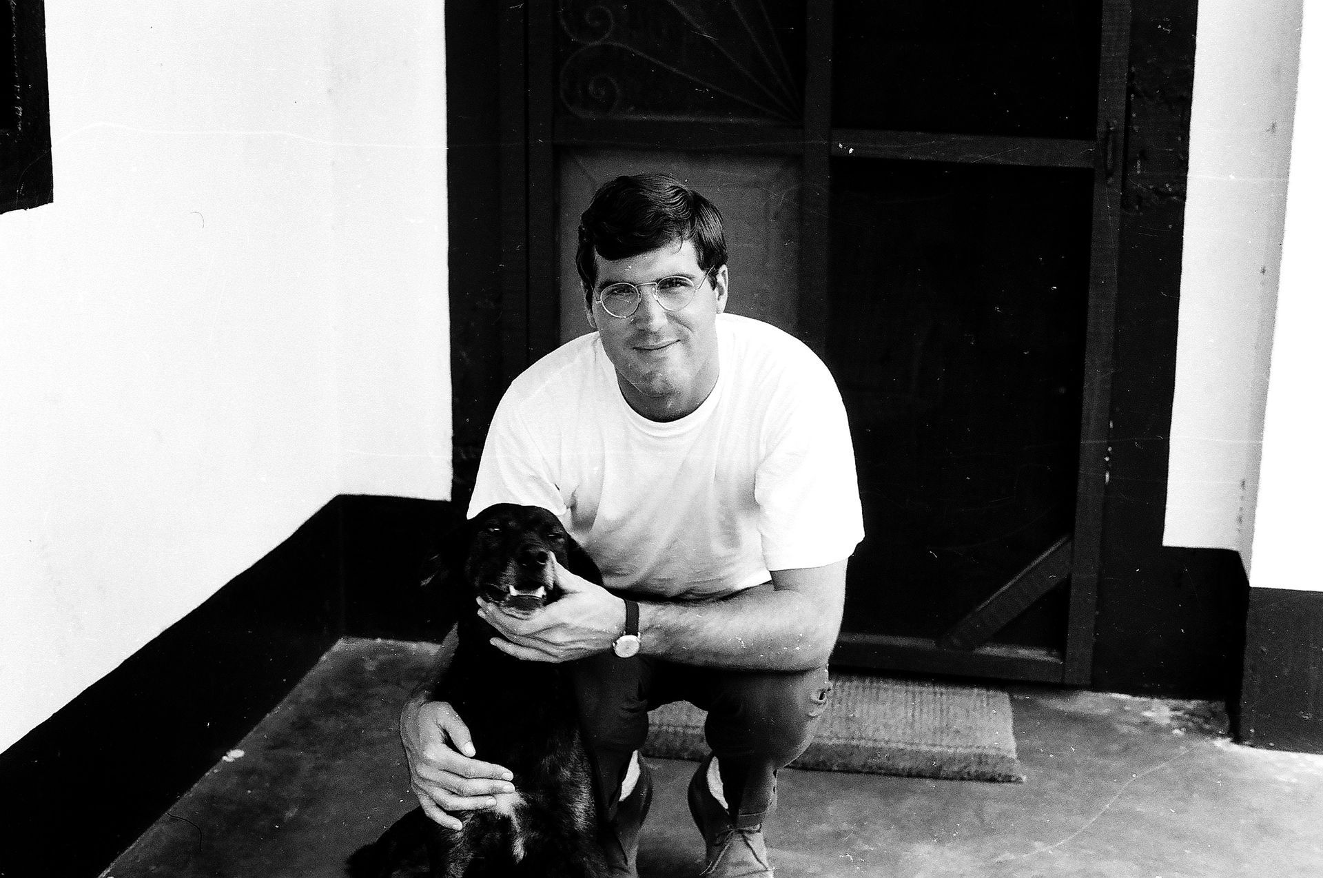 A man in a white shirt is kneeling down next to a black dog