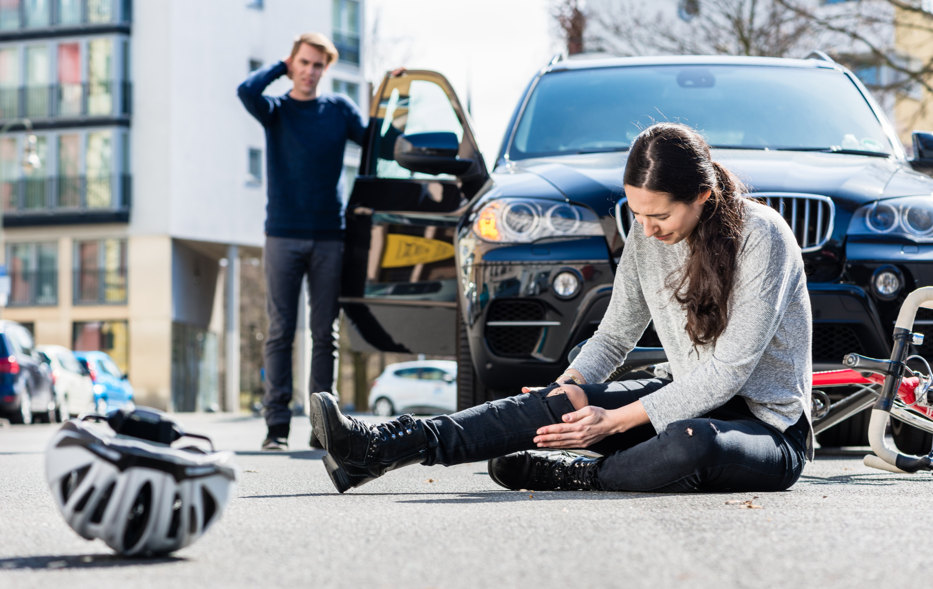 Woman injured on the ground after a collision with a car. Man stands behind, looking concerned. A helmet is nearby.