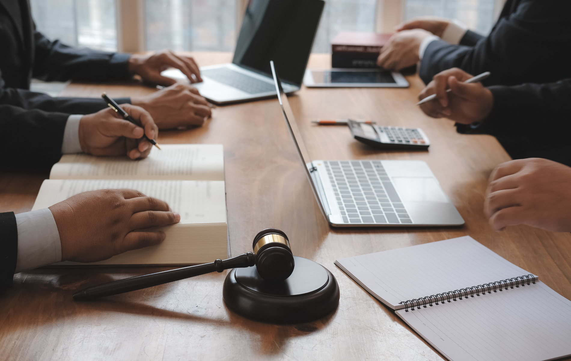 People in suits at a table with laptops, legal documents, and a gavel; they appear to be in a meeting.