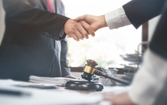 Two people shaking hands, gavel on a desk in the foreground, legal setting.