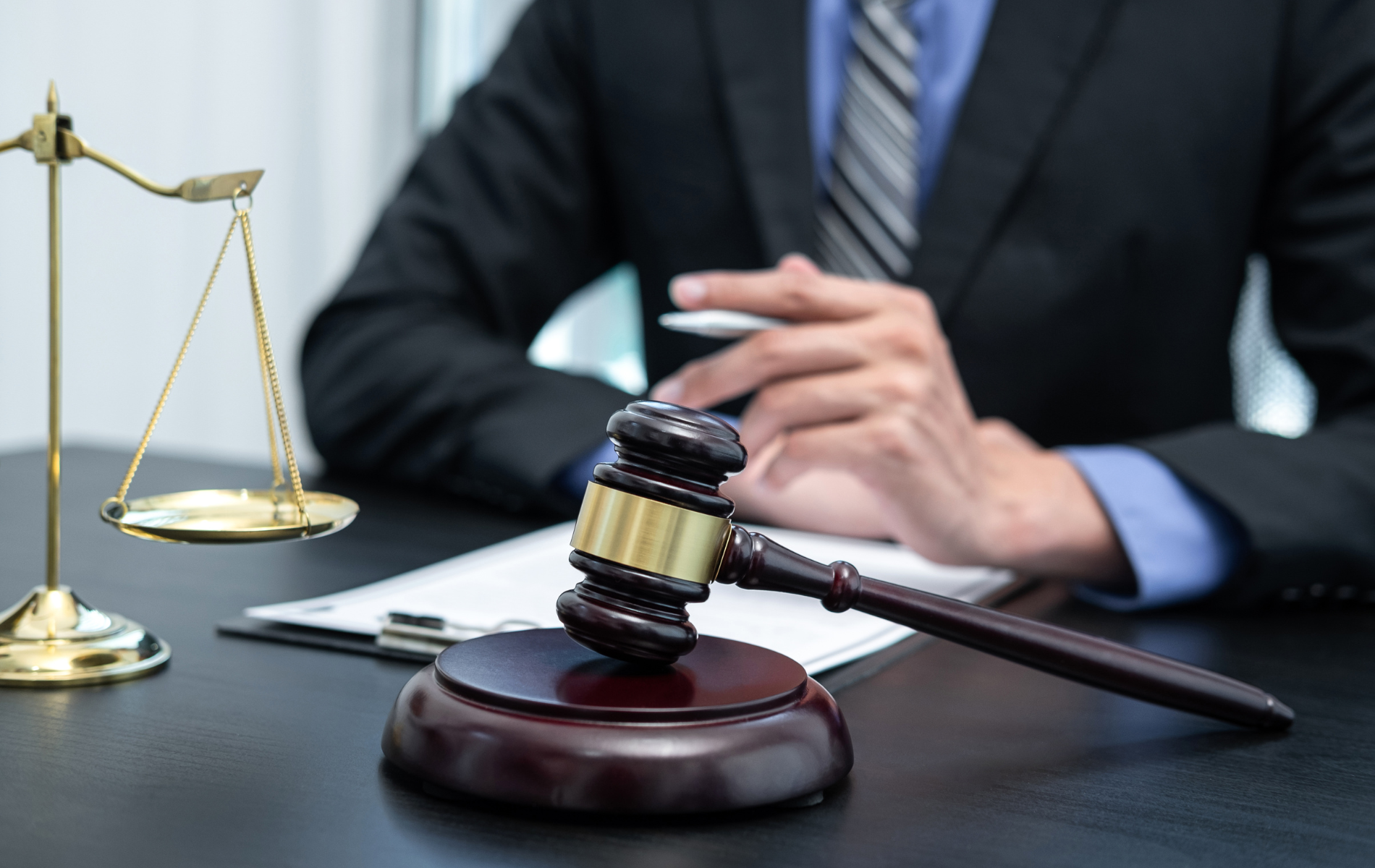 A gavel and scales of justice sit on a desk. A person in a suit is partially visible in the background.