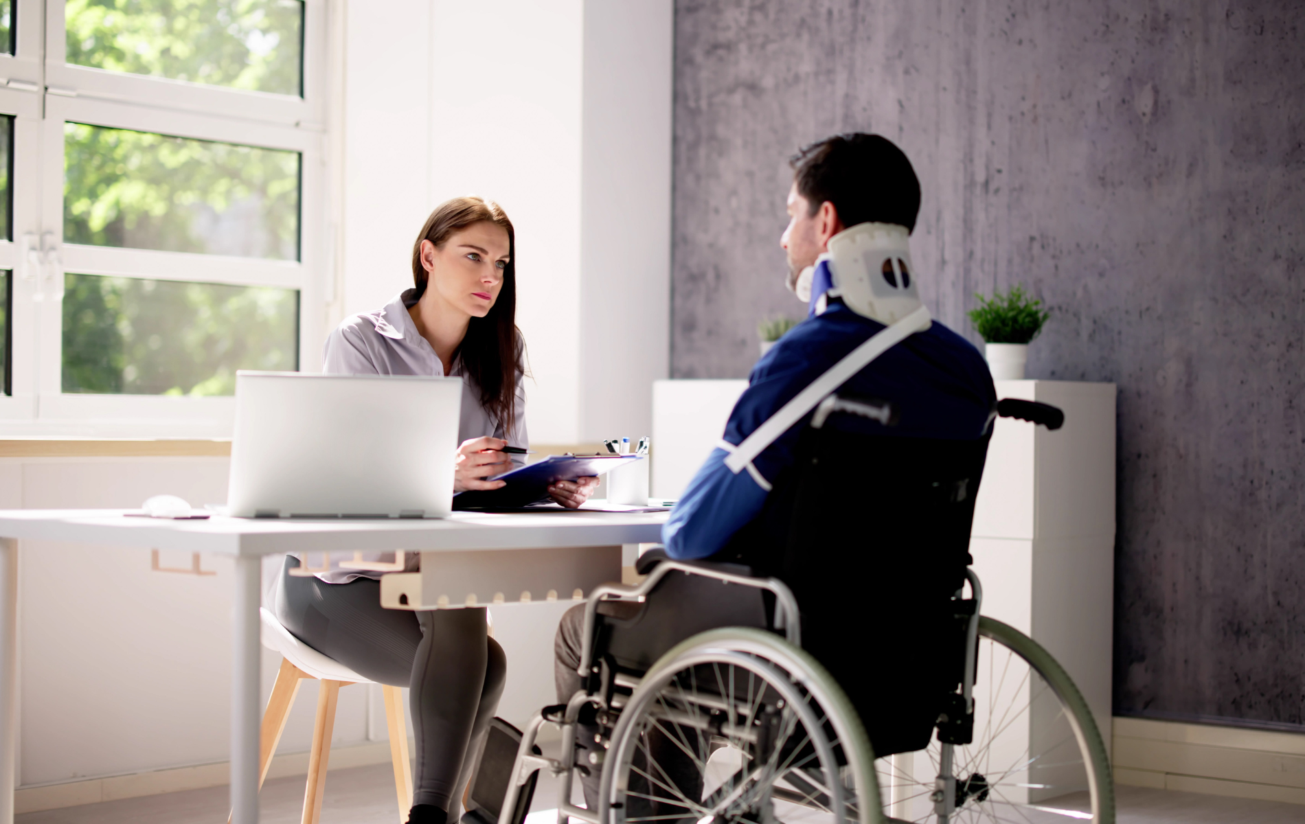 Woman interviewing person in wheelchair with neck brace. Office setting with laptop and window.