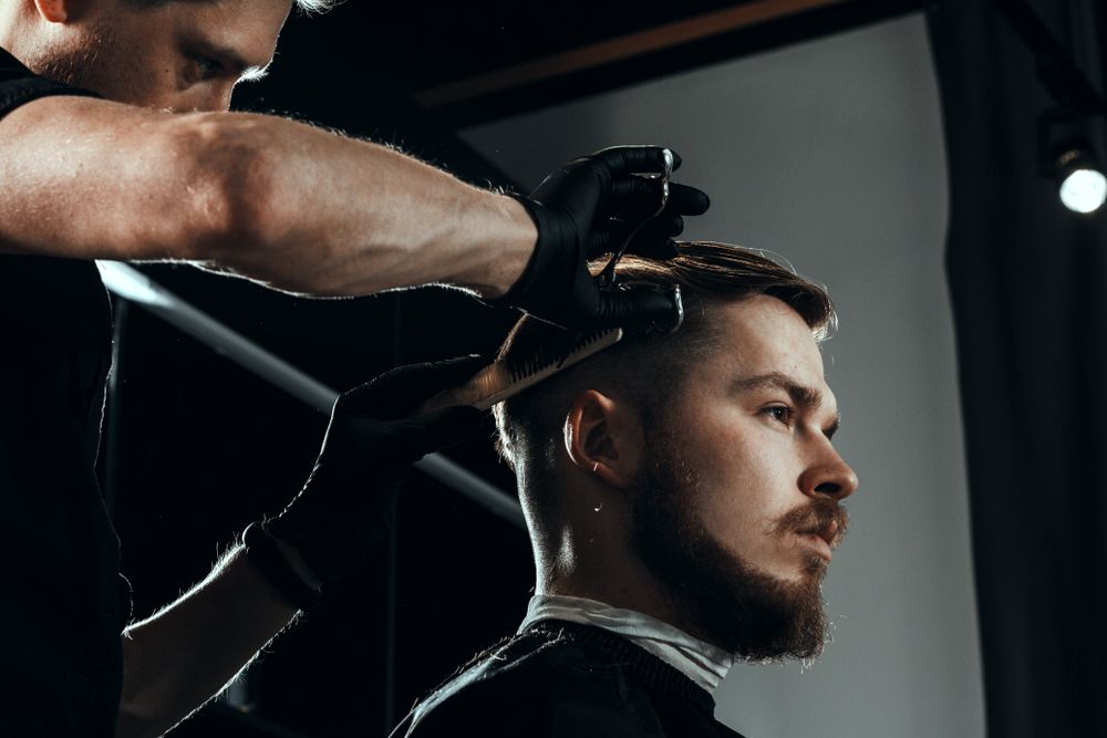 Barber cutting a client’s hair. Dark setting, the client has a beard. Barber wears black gloves.