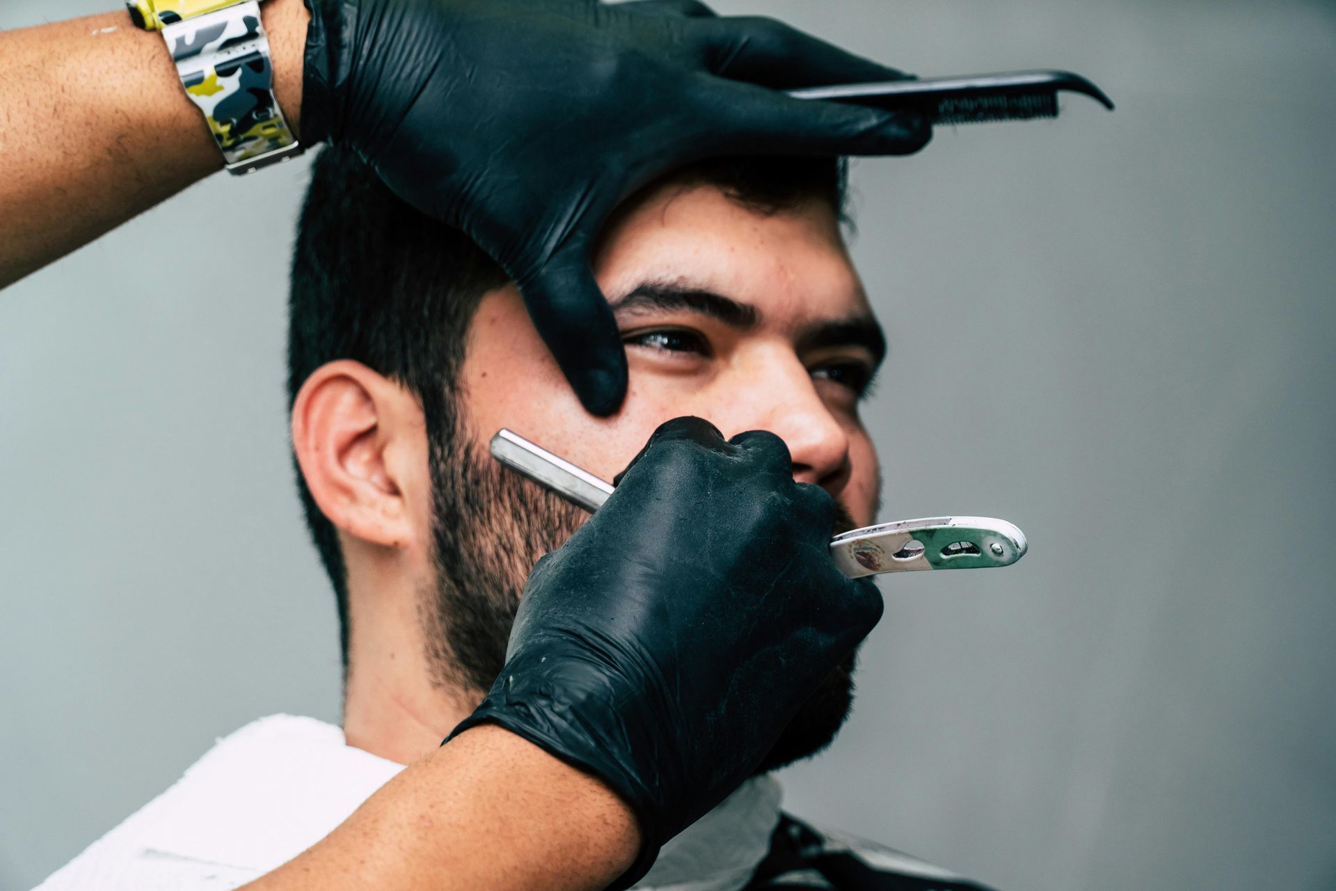 Barber shaving a man's beard with a straight razor. The barber is wearing black gloves.